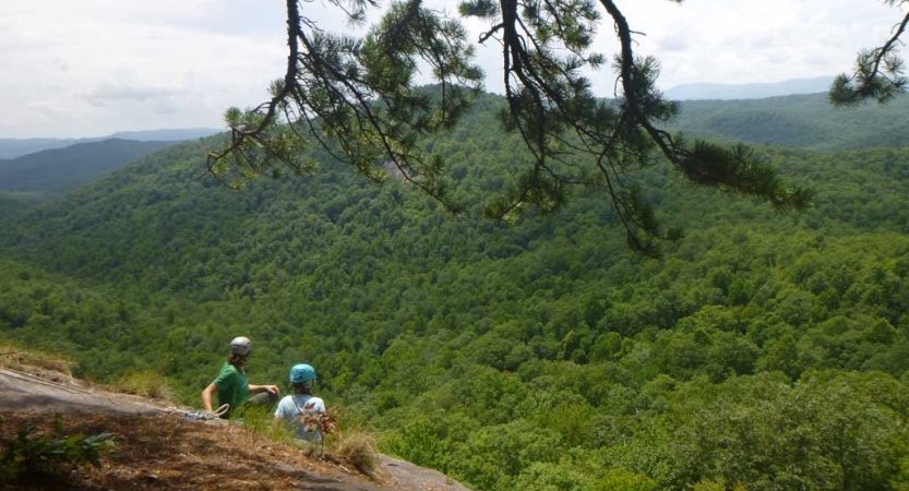 Two people wearing helmets sit on a rocky slope overlooking the vast forested area below. 